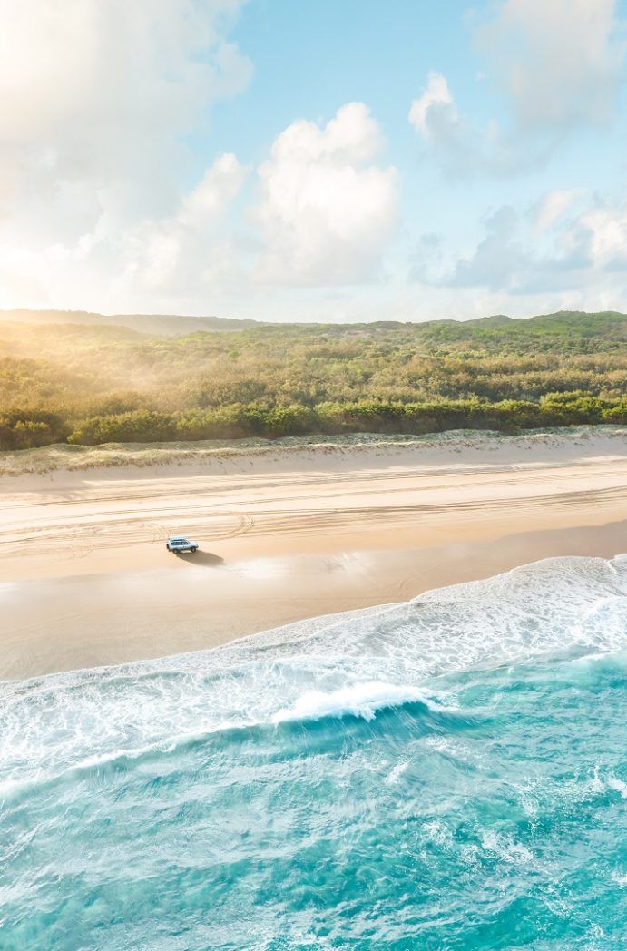 Vue aérienne d'une plage de Stradbroke Island avec un 4x4 roulant sur le sable, entourée d'eau turquoise et de végétation luxuriante