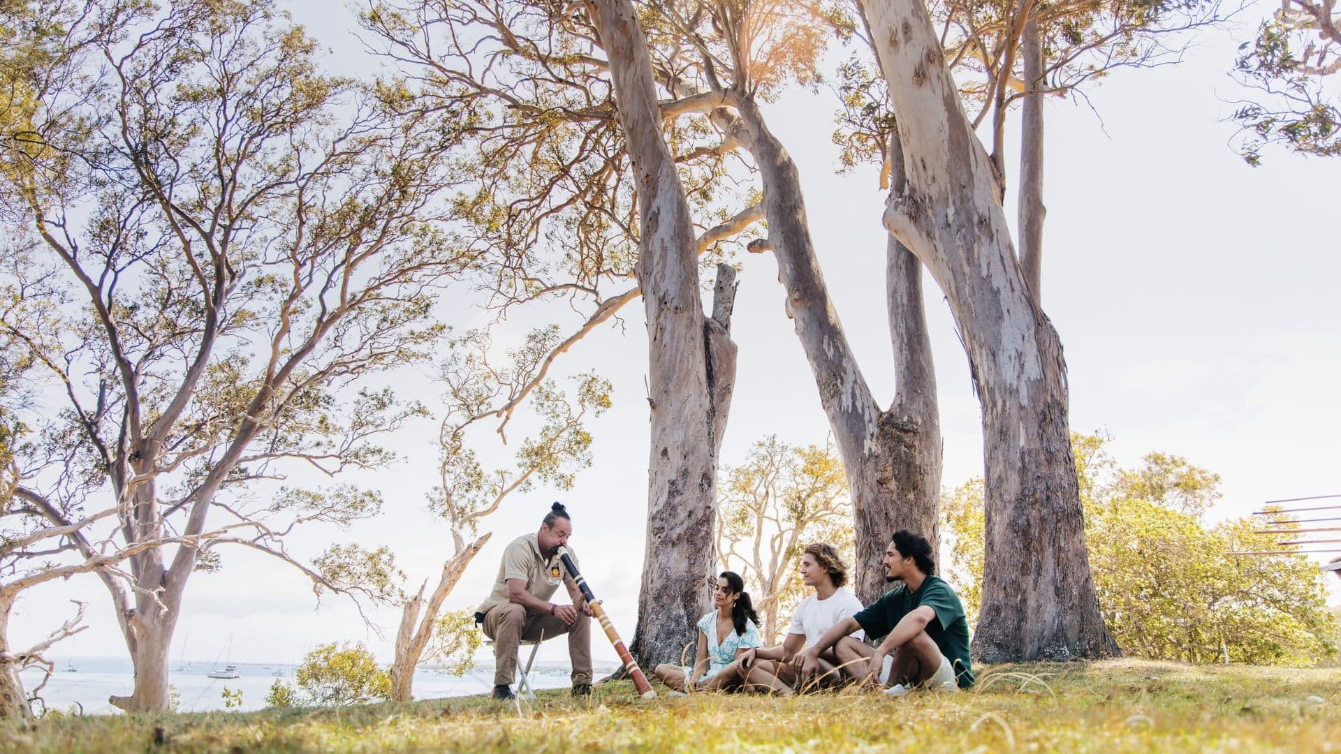 Quandamooka man Matt Burns plays the didgeridoo for a small tour group seated beneath gum trees on North Stradbroke Island (Minjerribah)