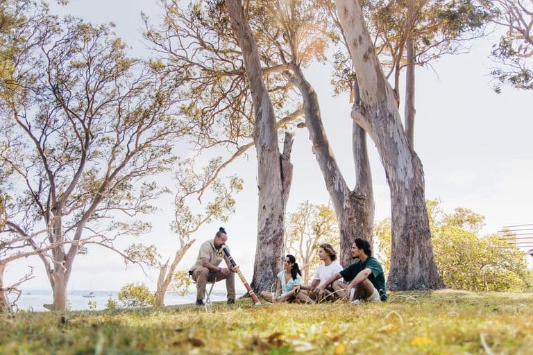 Quandamooka man Matt Burns plays the didgeridoo for a small tour group seated beneath gum trees on North Stradbroke Island (Minjerribah)